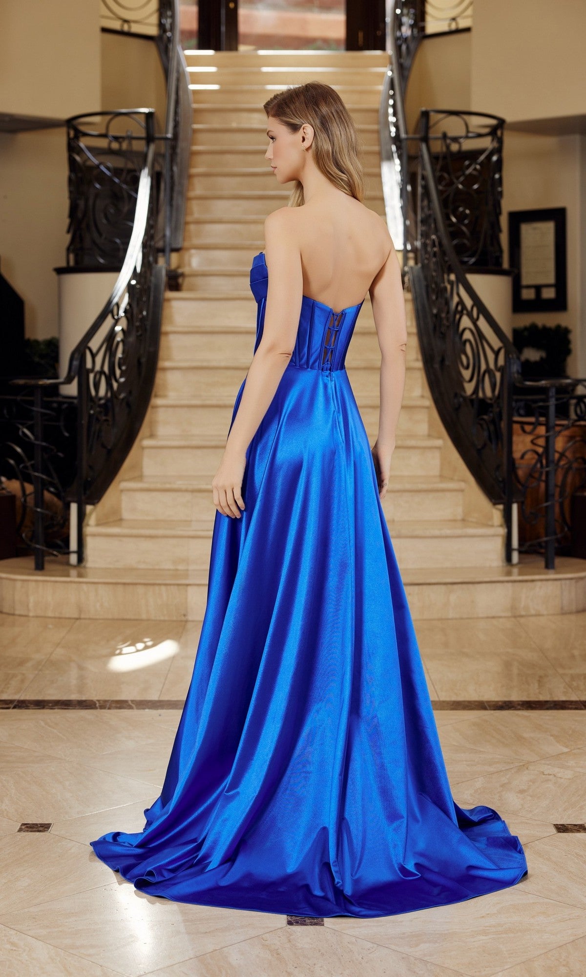 A woman stands at the base of a staircase in the Nox Anabel K1735 Long Prom Dress—a strapless satin gown featuring an elegant corset bodice, laced back detail, and a royal blue A-line floor-length skirt.